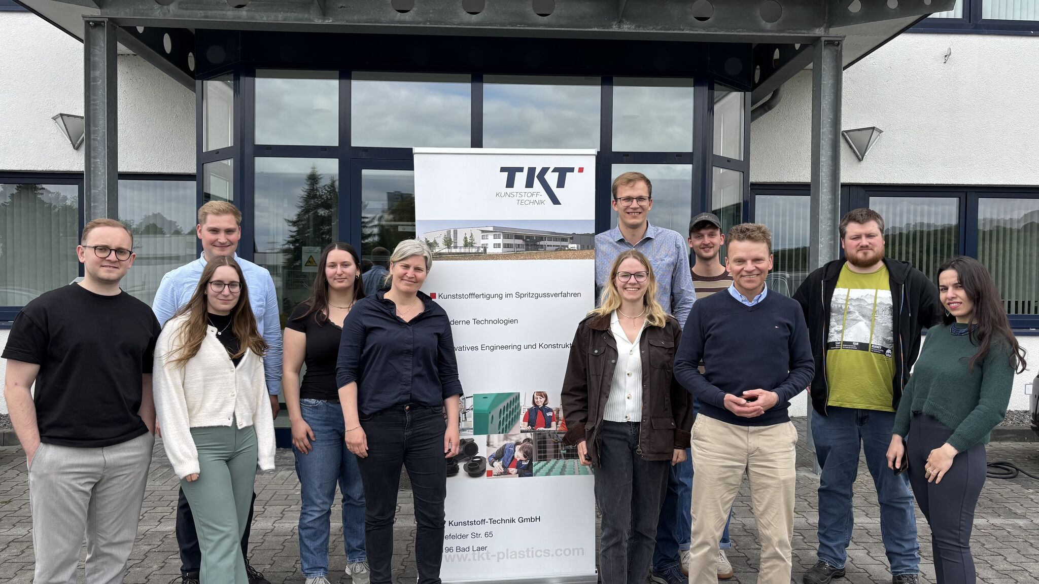 Eine Gruppe von 11 Personen posiert lächelnd vor einem Gebäude mit einem Banner von "TKT Kunststoff-Technik".