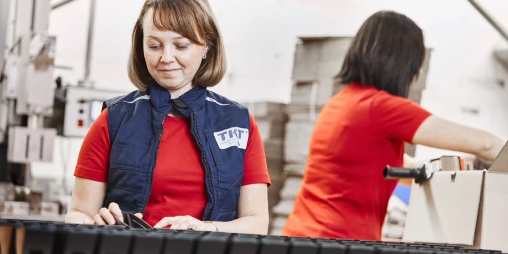 Zwei Frauen arbeiten konzentriert in einer Fabrikumgebung, eine mit blauer Weste, die andere mit Karton.