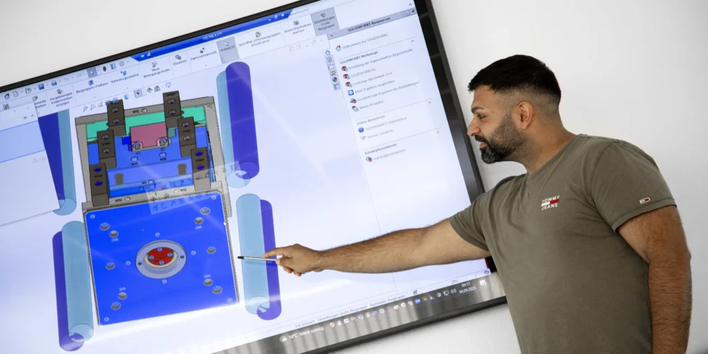 Eine Person zeigt auf einen Bildschirm mit einer CAD-Zeichnung eines mechanischen Bauteils in SOLIDWORKS.