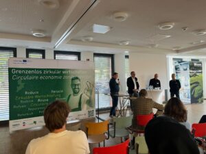 Ein Konferenzredner spricht über Kreislaufwirtschaft, während ein Banner Nachhaltigkeitsbegriffe zeigt.