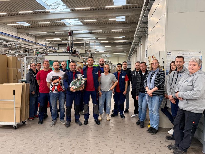 Eine Gruppe von Menschen posiert in einer Fabrik, einige in Arbeitskleidung, mit einem Präsentkorb.