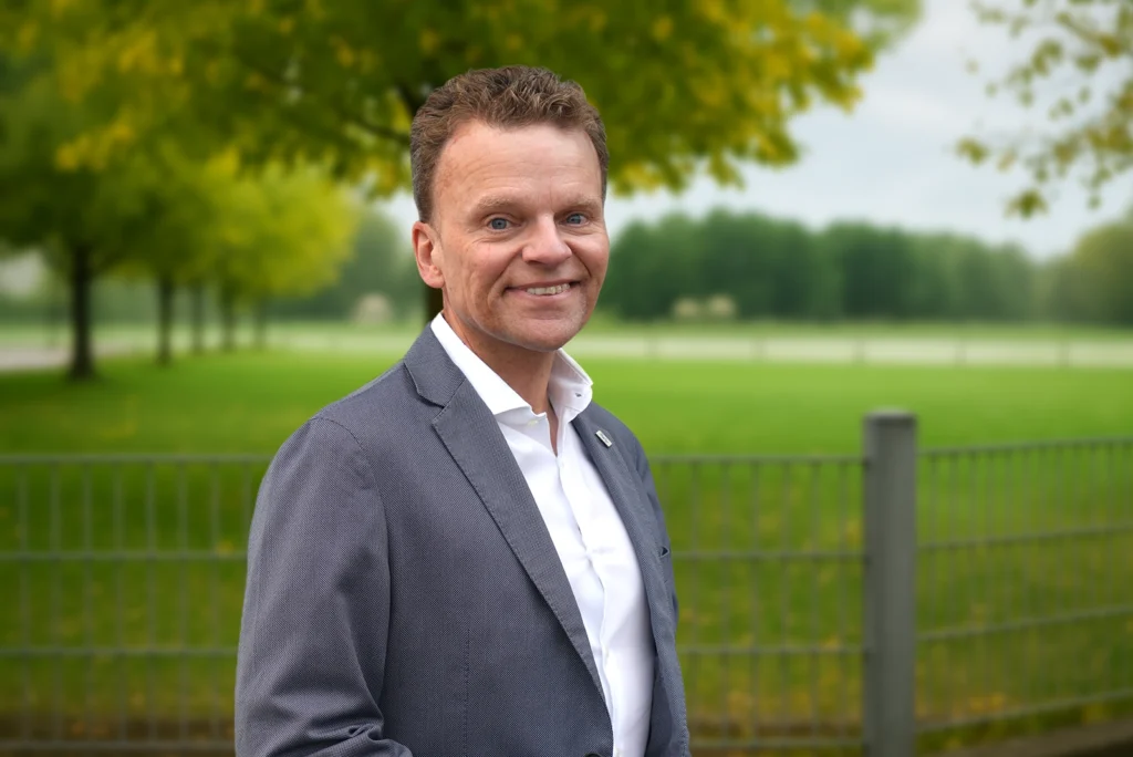 Person im grauen Anzug vor unscharfem, grünem Hintergrund; freundlich und professionell wirkend.