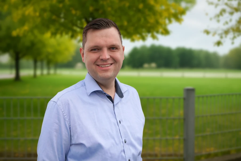 Eine lächelnde Person im hellblauen Hemd steht vor einer unscharfen grünen Wiese mit Bäumen.