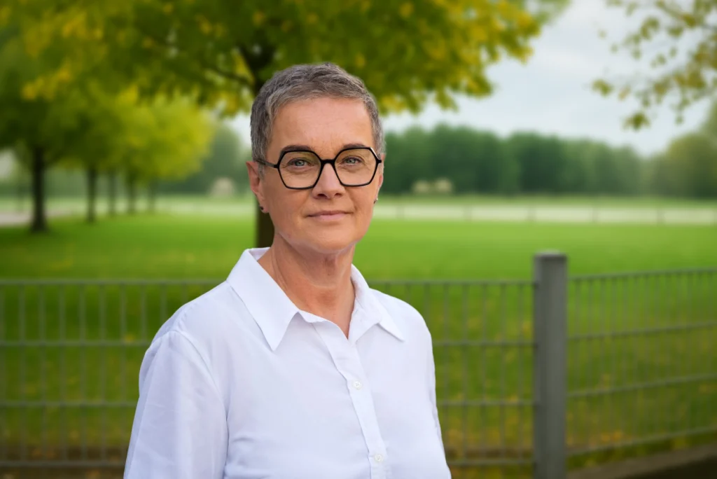 Eine Person mit Brille und weißem Hemd steht in einem grünen Park und schaut direkt in die Kamera.