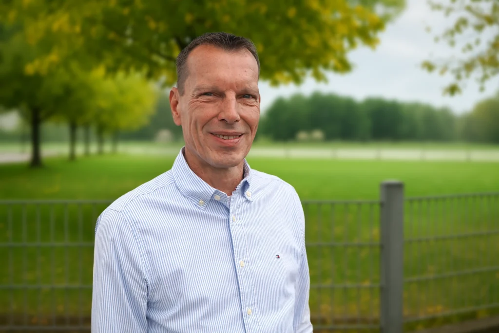 Ein lächelnder Mann im gestreiften Hemd steht vor einer grünen Wiese mit Bäumen im Hintergrund.