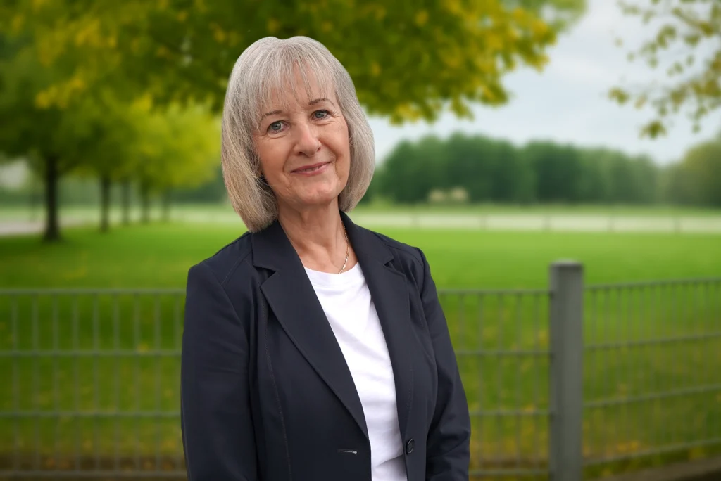 Eine lächelnde Frau im Blazer steht vor einem grünen, parkähnlichen Hintergrund mit Bäumen und Zaun.